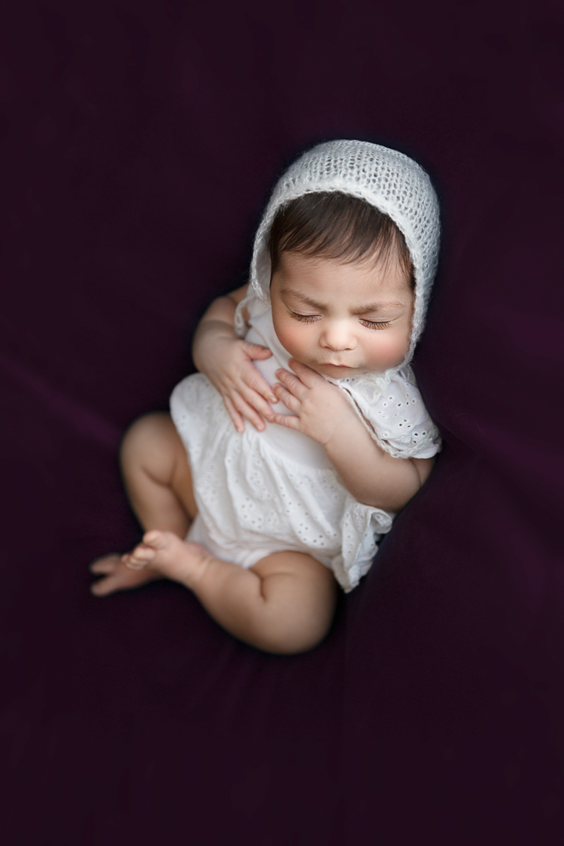 A sweet newborn girl is posed on a purple velvet background in a delicate white eyelet gown and white knitted baby bonnet. The image illustrates the quality of photography you can expect from Heather Tristan of Laughing Magpies Photography