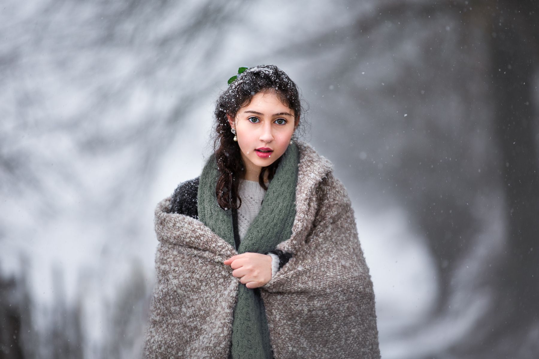 An award-winning teen portrait by Carthage, North Carolina photographer Heather Tristan for Laughing Magpies Photography. The image shows a young teen girl dressed warmly in a thick woolen sweater, coat, and scarf, and wrapped in a rustic-textured blanket. She stands alone on a snowy lane as the snow falls softly on and around her, collecting in white drifts on her head and shoulders. A monochromatic tree behind her and to her left creates a tunnel-like space that creates an even more mystical feeling for this magical snow portrait.